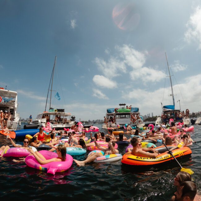 A large group of people enjoying a sunny day on the water, floating on colorful inflatable rafts and tubes. Several boats from The Yacht Social Club Sydney Boat Hire are anchored nearby with more people on board, and the sky is partly cloudy. The atmosphere is festive and lively.