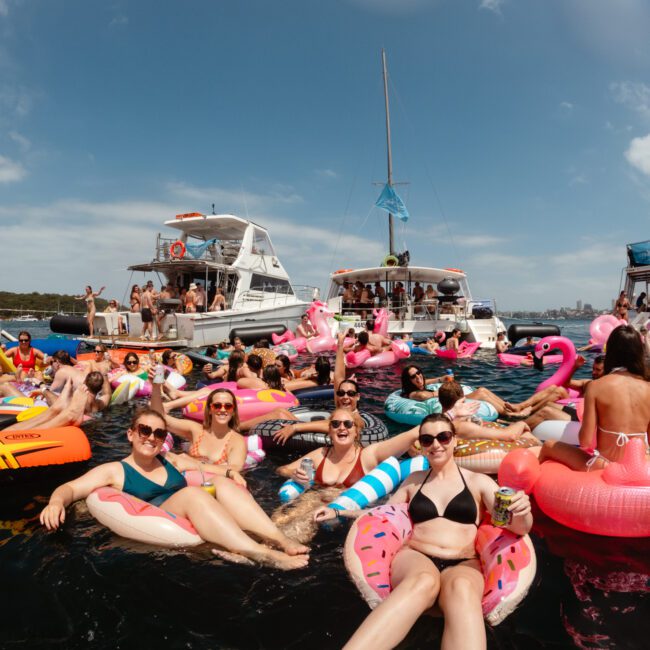 A lively scene on the water with a large group of people relaxing on colorful inflatable floats, including doughnuts, unicorns, and flamingos. Boats from Sydney Harbour Boat Hire are anchored in the background as everyone enjoys a sunny day on the water.