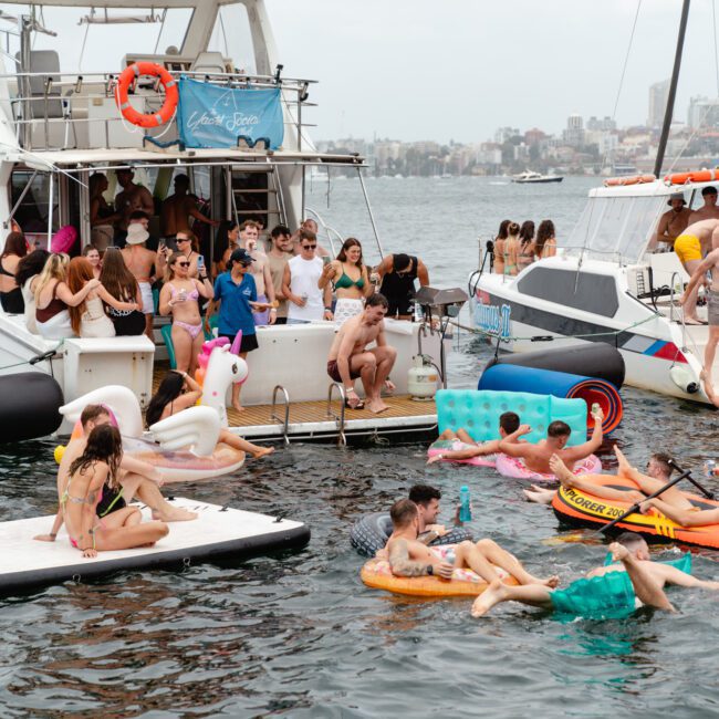 People are having a lively time on and around boats anchored in a body of water. Some are lounging on colorful inflatables, including a unicorn float, while others swim or relax on paddleboards. The sky is overcast, and a city skyline is visible in the distance, setting the scene for The Yacht Social Club Event Boat Charters.
