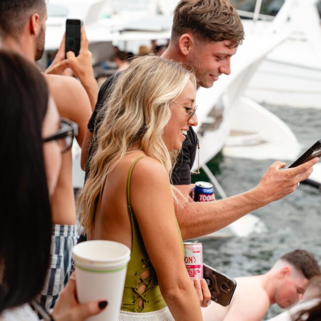 A lively group of people enjoying a sunny day on a luxury yacht. The focus is on a woman in a green top and white skirt, holding a drink, smiling at a man beside her who is also holding a drink and looking at his phone. Several other people are in the background, some taking photos during The Yacht Social Club event.