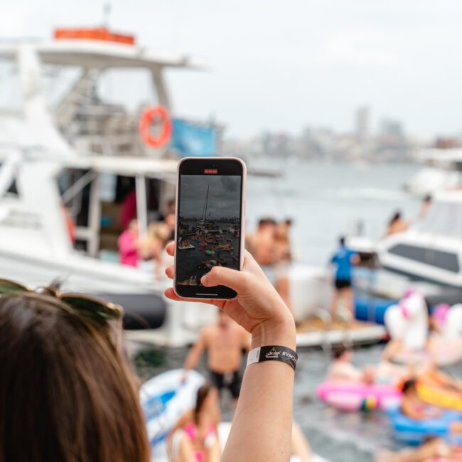 A person takes a photo with a smartphone at a lively beach party. The scene features partygoers on inflatable floats in the water and The Yacht Social Club Event Boat Charters in the background, creating an atmosphere of fun and relaxation under a cloudy sky.
