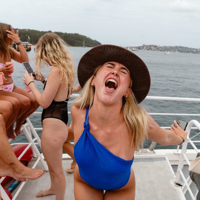 A woman in a blue one-shoulder swimsuit and a wide-brimmed hat joyfully leans forward, shouting or laughing. She is on The Yacht Social Club Event with friends, with water and a distant shoreline in the background. Other women in swimsuits are seated and standing around her, enjoying the sunny day.