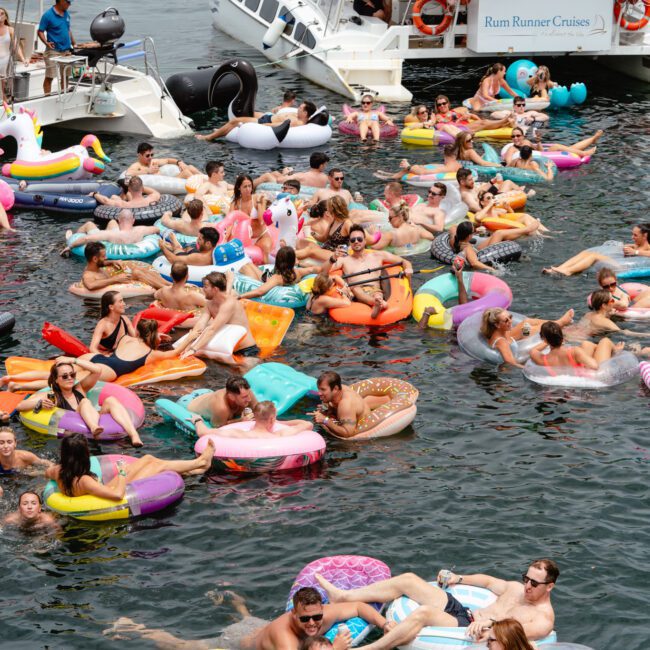 A crowded water party features numerous people on colorful inflatable tubes and rafts in a lake. Two boats, labeled "Rum Runner Cruises," are docked nearby with participants mingling and enjoying the sun. Everyone appears to be having a lively time at The Yacht Social Club event.