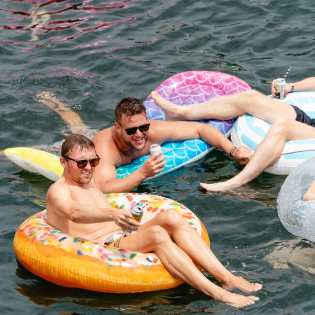 Two men relax and smile on inflatable floats in a body of water. One sits on a doughnut-shaped float, the other on a light blue float, both holding drinks. Other people in various colorful inflatables are seen in the background, reminiscent of fun Sydney Harbour boat parties with The Yacht Social Club.