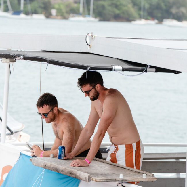 Two bearded men are shirtless, standing on a boat from The Yacht Social Club. One holds a can while using a phone, and the other leans forward on a table. Both wear sunglasses and swim trunks. In the background are other boats and a tree-lined shoreline.