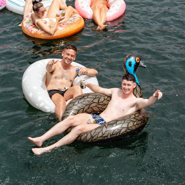 A group of people enjoy floating on inflatable rings in a body of water. Two men in the foreground smile and gesture to the camera, with one wearing a playful bird-shaped hat. Others relax on various colorful inflatables in the background, holding drinks and enjoying the sun, as part of The Yacht Social Club Event.