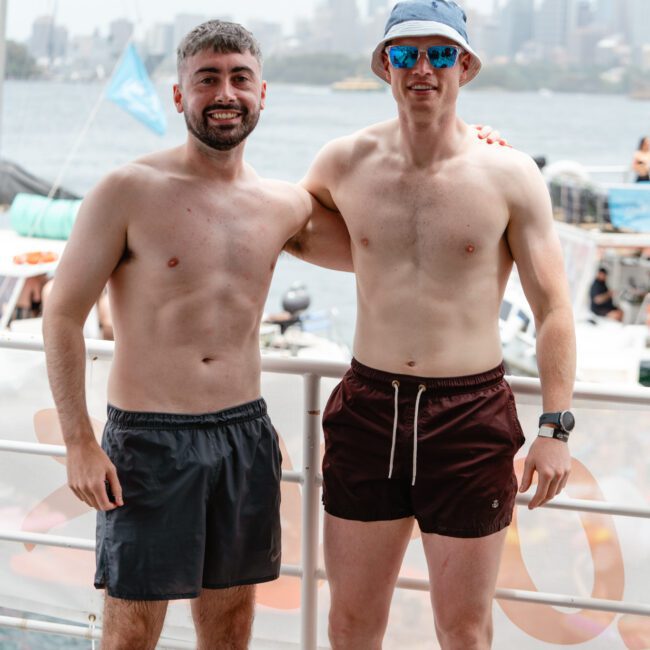 Two men in swim trunks, one in gray and the other in dark red, stand side by side with their arms around each other. Both are shirtless and smiling. The man on the right wears a blue bucket hat and sunglasses. A body of water and cityscape are visible in the background, suggesting a fun day at The Yacht Social Club Sydney Boat Hire.