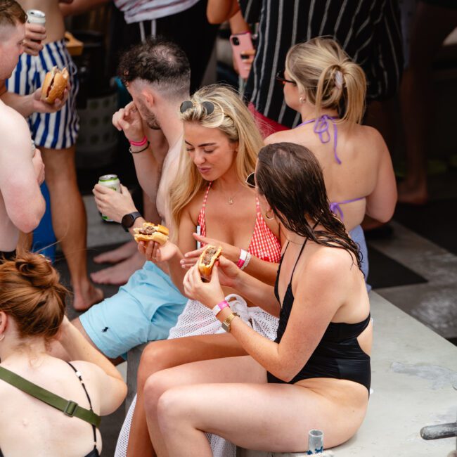 A group of people in swimsuits are gathered by a pool at The Yacht Social Club Event. Two women are seated, eating hot dogs and engaged in conversation, while others stand or sit nearby, socializing and holding drinks. A mix of summer attire and poolside activity reflects the vibrant atmosphere.