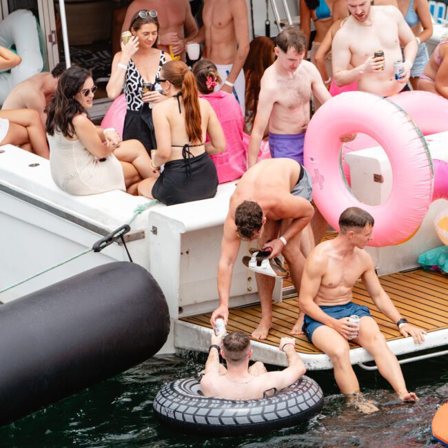 A group of people on a boat enjoying a sunny day. Some are sitting on the deck, while others are in the water with inflatables, including a tire tube and a pink flamingo. One person is handing a beverage to another in the water. Everyone appears relaxed and happy, making it an ideal scene for The Yacht Social Club Sydney Boat Hire.