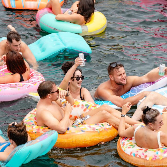 A group of people relax and socialize on colorful inflatable pool floats in a body of water. Many are holding drinks and wearing sunglasses. The floats resemble donuts, pretzels, and other fun shapes, creating a festive atmosphere akin to parties by The Yacht Social Club Event Boat Charters.