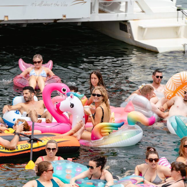 A lively group of people enjoying a sunny day on the water. They are floating on various colorful inflatable pool floats, including flamingos, unicorns, and boats, near a docked vessel from The Yacht Social Club Sydney Boat Hire. Everyone appears to be relaxed and having a great time.