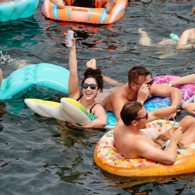 A group of people enjoying a summer day on a lake, relaxing on colorful inflatable floats. One person raises a can in celebration while others smile and chat. The floats, shaped like donuts, pizza slices, and lounge chairs, create a lively atmosphere reminiscent of The Yacht Social Club's luxury yacht events in Sydney.