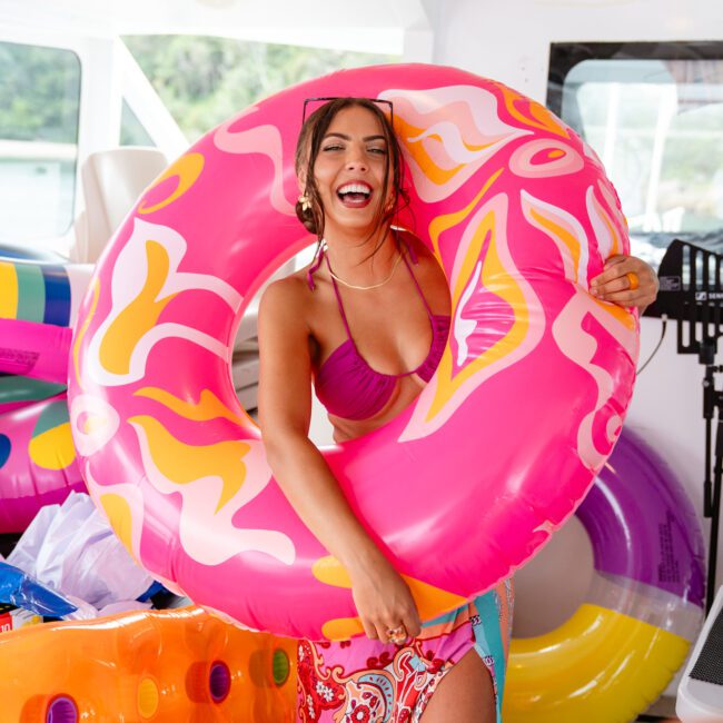 A woman on a boat smiles broadly while holding a large, colorful inflatable ring. She wears a vibrant pink bikini top and a colorful, patterned sarong. More inflatables and boating gear are visible around her, setting the stage for The Yacht Social Club Sydney Boat Hire's lively and summery events.