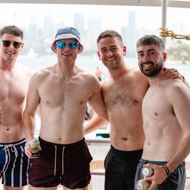 Four men in swimwear stand together on a boat, smiling at the camera. They are holding drinks with a city skyline visible in the background. Enjoying a sunny day out on the water, it’s clear they’re participating in one of The Yacht Social Club Event Boat Charters.