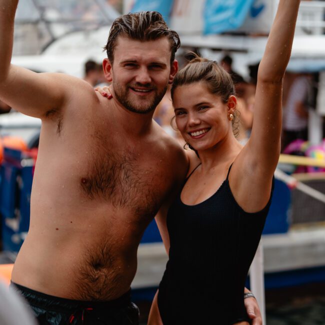 A man and a woman, both in swimwear, smile and wave at the camera while standing on a dock with boats in the background. The man is shirtless and wears dark swim trunks, and the woman is in a black one-piece swimsuit. Enjoy events like these with Boat Rental and Parties Sydney The Yacht Social Club.