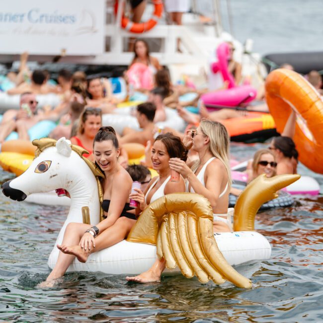 A group of people are enjoying a sunny day on water, many on inflatable floats. Three women are riding together on a large inflatable unicorn float, smiling and holding drinks. In the background, a Luxury Yacht Rentals Sydney vessel hosts more revelers from The Yacht Social Club Event Boat Charters.