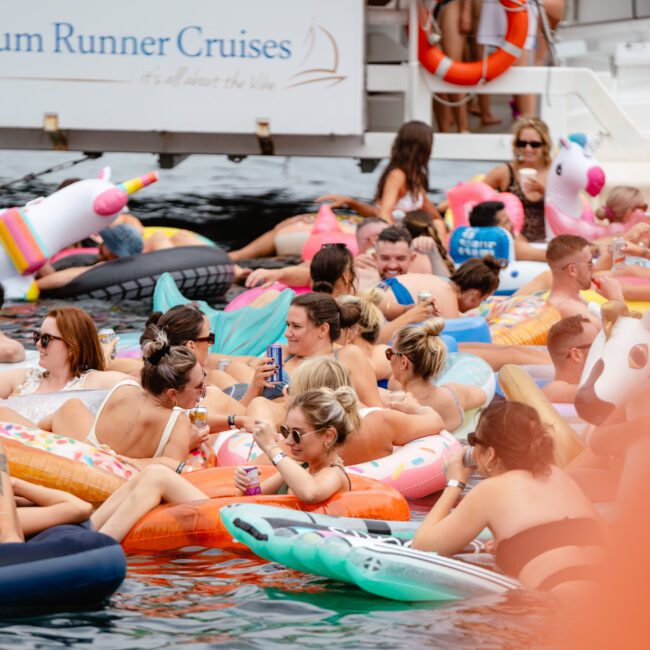 A lively crowd relaxes on inflatable floats of various shapes and colors, including unicorns, flamingos, and donuts, in the water near a cruise boat labeled "Rum Runner Cruises." The atmosphere at The Yacht Social Club is festive with everyone appearing to enjoy themselves.