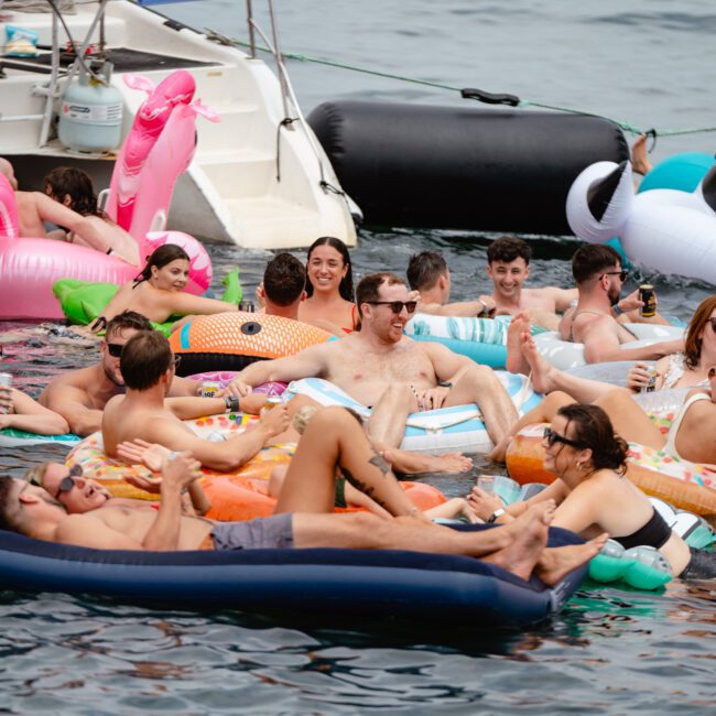 People are lounging on colorful inflatable floats in the water near a boat, socializing and enjoying drinks. Various float shapes include flamingos, unicorns, and pizzas, creating a lively and festive atmosphere. Experience this at The Yacht Social Club Sydney Boat Hire for unforgettable boat parties.