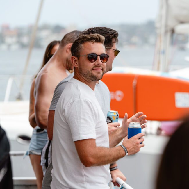 A man wearing a white t-shirt and dark sunglasses holds a drink while standing on a boat at The Yacht Social Club Sydney Boat Hire event. Other people, some in swimwear, are around him, and the background shows a blurred coastal scenery with buildings and water.