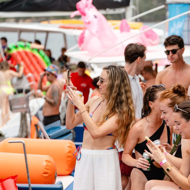 A group of people enjoys a boat party with various inflatables, including a pink unicorn. A long-haired woman in white pants and a bikini top takes a photo with her phone while others socialize around her. The Yacht Social Club Event Boat Charters offers an unforgettable experience, with a scenic bridge in the background.