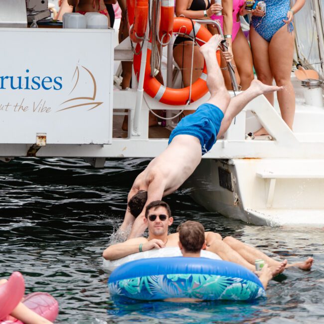 A man in blue swim trunks jumps off a boat into the water where other people are swimming and lounging on colorful inflatable rings. The boat, part of The Yacht Social Club Event Boat Charters, has a sign saying "Dinner Cruises," and people onboard are wearing swimsuits and life jackets. The atmosphere is lively.