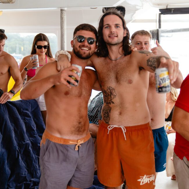 Two shirtless men are smiling and holding beer bottles while posing on a boat at The Yacht Social Club Event. The man on the left is wearing sunglasses and blue swim shorts, while the man on the right has long hair, a tattoo on his arm, and is wearing orange swim shorts. Other people are in the background.