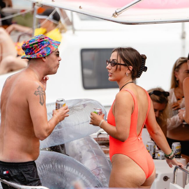 Two people are standing on a boat, conversing and holding drinks. The man on the left wears a colorful hat and sunglasses, has tattoos, and is shirtless. The woman on the right wears an orange one-piece swimsuit and sunglasses. Inflatable floats are visible in the background at The Yacht Social Club Event Boat Charters.