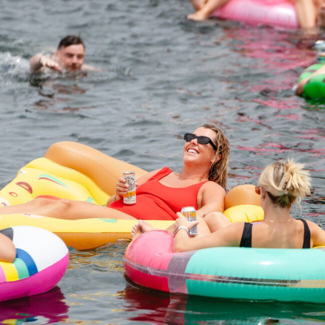 People are floating in a body of water on colorful inflatable pool floats. A woman in a red swimsuit is lying on a yellow float holding a can, smiling and wearing sunglasses. Nearby, others enjoy the water on various floats, with The Yacht Social Club Event Boat Charters adding to the fun atmosphere.