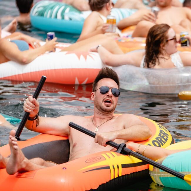 A group of people are floating on inflatable rafts and tubes in the water. The foreground shows a man wearing sunglasses relaxing in an orange inflatable raft, holding paddles. Other individuals are seen in the background, enjoying the sunny outdoor setting, reminiscent of boat parties by The Yacht Social Club.