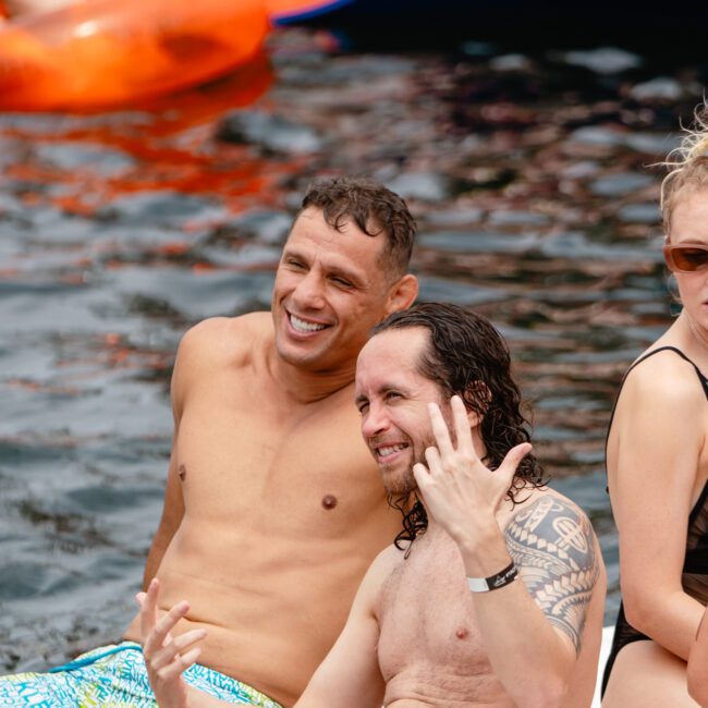 Two men are sitting on the edge of a pool, both smiling and laughing. The man on the left is wearing bright patterned swim trunks, and the man on the right, with long hair and tattoos on his arm, is wearing dark swim trunks. They appear to be having a great time, reminiscent of a gathering at The Yacht Social Club Sydney Boat Hire.