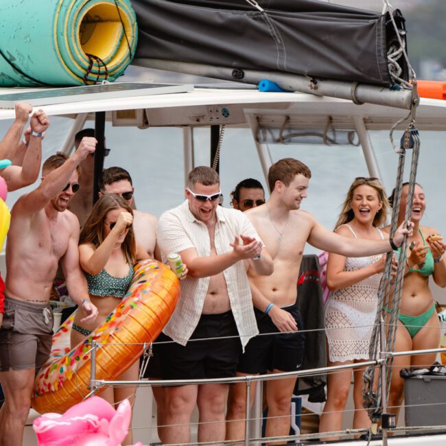 A group of people on a boat enjoying a sunny day. They are wearing swimsuits, holding inflatable pool toys, and cheering excitedly. The Yacht Social Club Sydney Boat Hire ensures everyone is smiling and having fun against the backdrop of a beautiful hilly coast.
