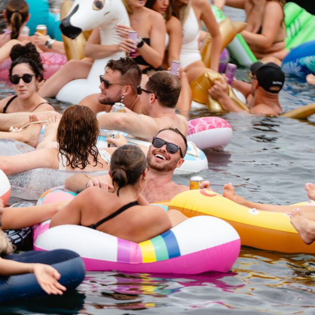 A large group of people floats on inflatable pool toys, including a unicorn and a duck, in a body of water. Many are wearing swimwear and sunglasses, smiling and enjoying themselves. The atmosphere is lively and festive, reminiscent of the vibrant gatherings hosted by The Yacht Social Club Sydney Boat Hire.
