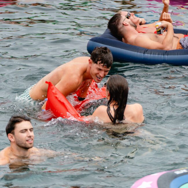 A group of people enjoying a day in the water. One man and one woman playfully splash each other. Another man relaxes on an inflatable floaty, while a fourth person swims nearby. All appear to be having fun, representing the lively spirit of Sydney Harbour Boat Hire The Yacht Social Club.