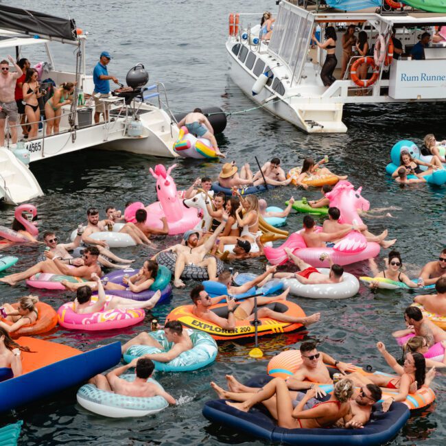 A lively scene of people on various inflatable floaties, including unicorns and donuts, gathered near several boats on a body of water. They're enjoying the day, socializing, and having fun in their colorful rafts. The weather appears pleasant and bright—a typical day with The Yacht Social Club Sydney Boat Hire.