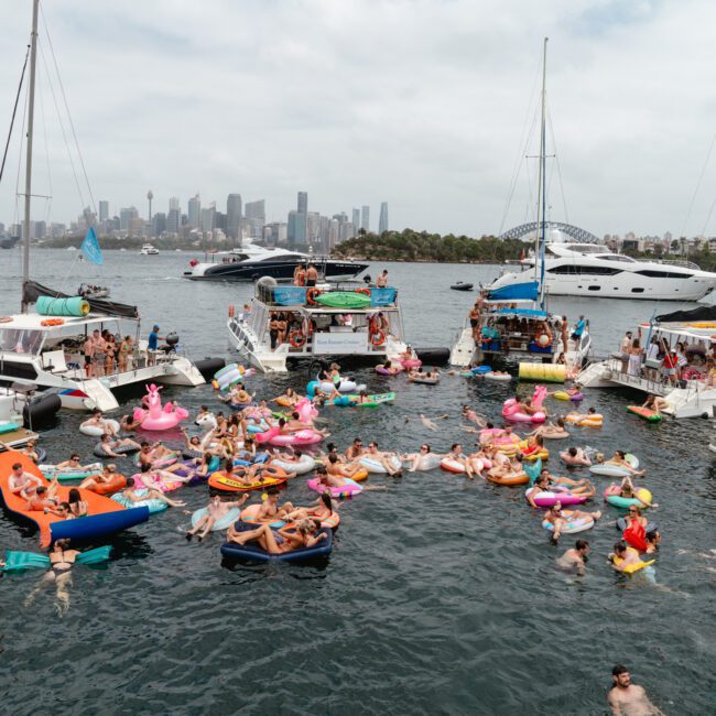 A lively scene of a boat party on a sunny day with numerous people on yachts and colorful inflatables in the water. The background features a city skyline and recognizable landmarks on the horizon. The atmosphere is festive and vibrant, reminiscent of Boat Parties Sydney with The Yacht Social Club.