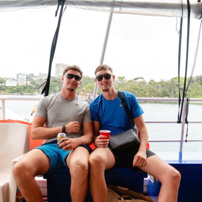 Two men sit closely side by side on a boat with their arms around each other, both wearing sunglasses and holding drinks. One wears a grey shirt and blue shorts, the other blue shirt and grey shorts. Other passengers and a coastline are visible in the background—capturing moments from The Yacht Social Club Event Boat Charters.