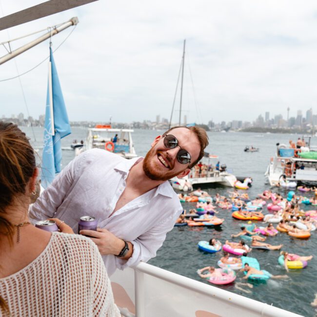 A man in a white shirt and sunglasses leans over the railing of a boat, smiling. A woman with a bun and a beige sweater stands nearby holding drinks. The background shows colorful floats and boats on the water under a cloudy sky, embodying the spirit of Boat Parties Sydney The Yacht Social Club.