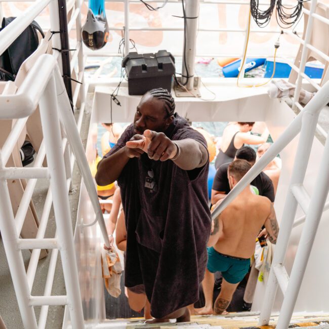 A man wearing a black towel gestures towards the camera while ascending the steps on a boat. Behind him, people in swimwear are gathered and a beach can be seen in the distance through the boat's railings. Equipment and life jackets hang on the walls around the stairs, depicting a lively Yacht Social Club event.