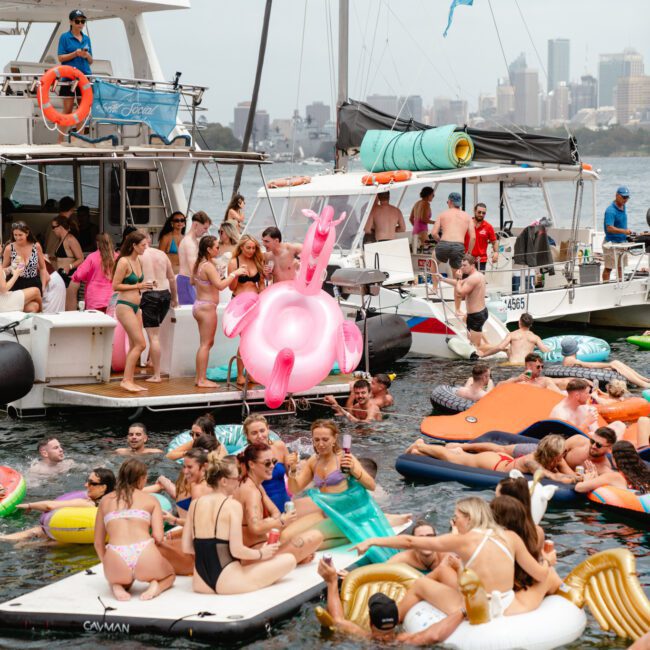 A lively scene of people enjoying inflatable floats and boats in the water, some drinking and socializing. The sky is overcast, with a city skyline visible in the background. Vibrant inflatables, including a large pink flamingo, add to the fun atmosphere. Consider Sydney Harbour Boat Hire The Yacht Social Club for your next adventure!