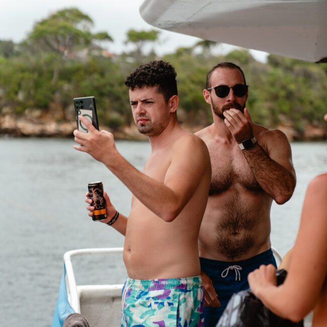 Two men stand on a boat deck by the railing, with water and trees in the background. One man is holding a drink and taking a selfie. The other man, shirtless and wearing sunglasses, enjoys a snack. They seem to be part of The Yacht Social Club Sydney Boat Hire event. Another person's shoulder is partially visible.