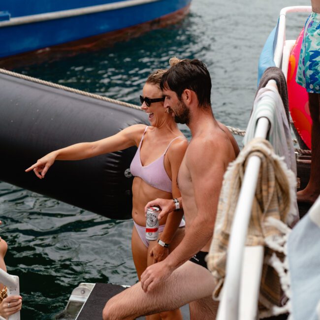 A woman and a man stand on the deck of a boat in swimsuits, with the woman pointing towards the water. They are smiling and appear to be enjoying a sunny day by the sea. A blue and white boat is visible in the background, likely part of The Yacht Social Club Sydney Boat Hire.