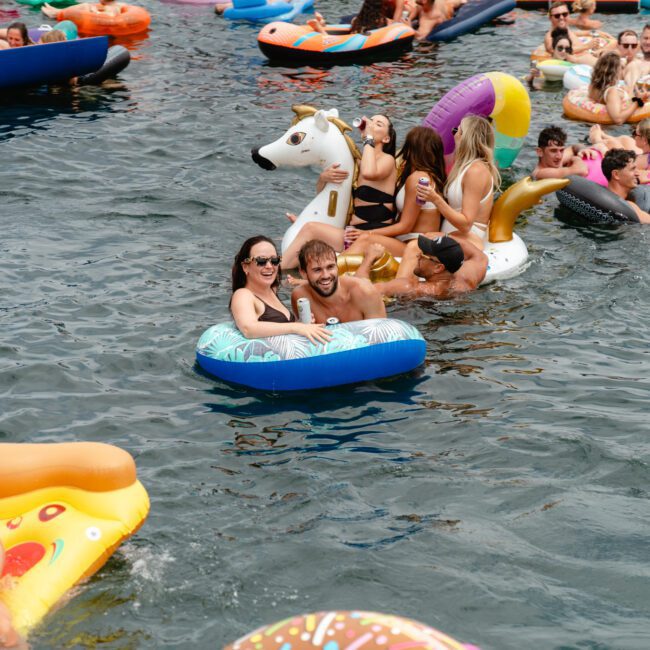 A lively scene with people enjoying a sunny day floating in a body of water on various inflatable devices, including a unicorn, a swan, and pizza slice. Hosted by The Yacht Social Club Sydney Boat Hire, they are smiling, laughing, and holding drinks, creating a fun and festive atmosphere.