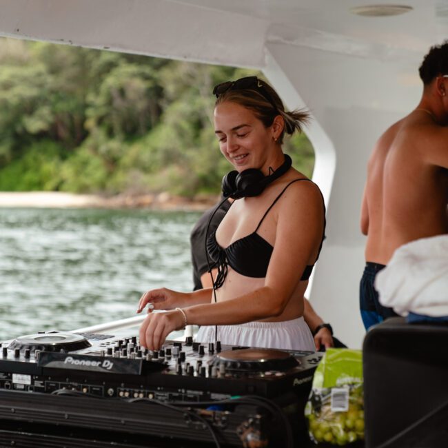 A woman in a black bikini top and white skirt is DJing on a boat, smiling as she works the turntables. She has headphones around her neck. The background shows a body of water and a green, tree-lined shore. Another person nearby in swim trunks adds to the lively atmosphere of The Yacht Social Club event by Luxury Yacht Rentals Sydney.