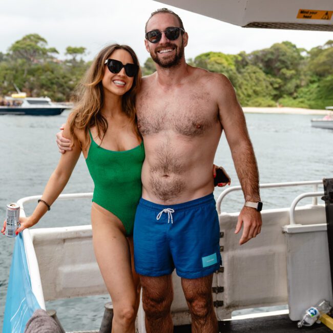 A woman in a green swimsuit and a man in blue swim trunks are smiling and standing close together on a boat. Both are wearing dark sunglasses. The background shows a serene water body with boats and lush greenery, perfect for enjoying Luxury Yacht Rentals Sydney through The Yacht Social Club.