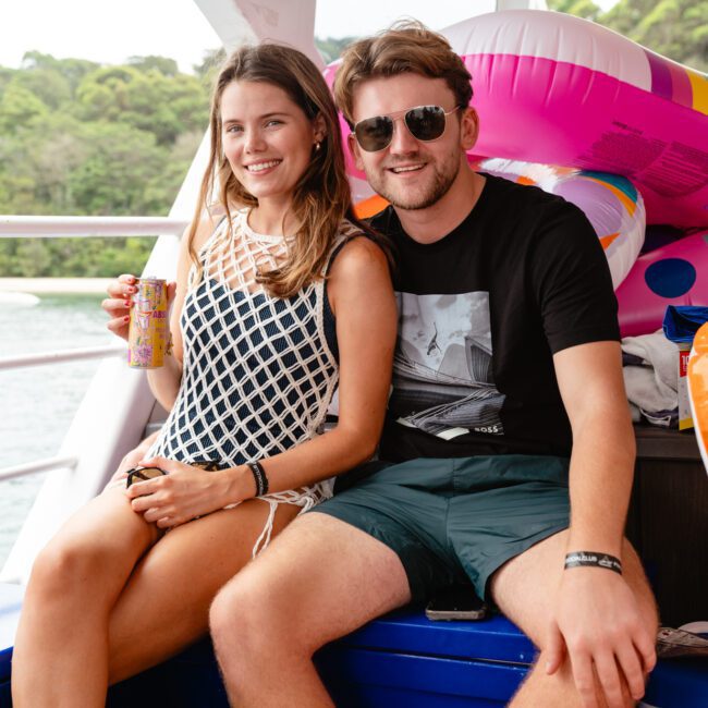 A woman and man smile while sitting together on a boat, enjoying a beverage and stylish sunglasses. They are surrounded by colorful inflatable pool toys. The background shows water and greenery, encapsulating the vibrant atmosphere of Boat Parties Sydney The Yacht Social Club.