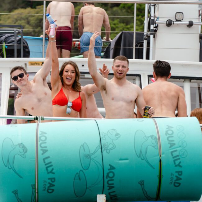 A group of six people in swimsuits is enjoying a sunny day on a boat. Three in the front, two men and one woman, are smiling and raising their hands. A large pool float labeled "Aqua Lily Pad" rests on the boat's side. Trees are visible in the background as they revel with The Yacht Social Club Sydney Boat Hire.