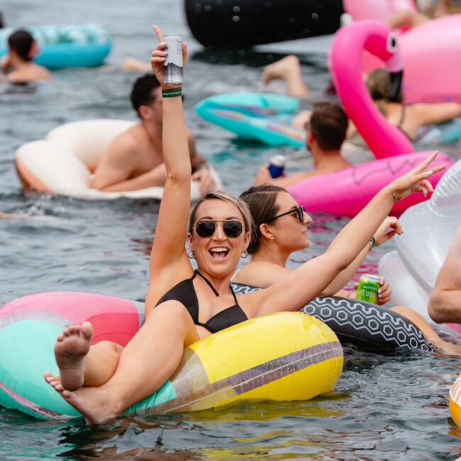 A group of people enjoying a fun time in the water on colorful inflatable floats. A woman in sunglasses and a black bikini sits on a pink and yellow float, smiling and giving a thumbs-up with one hand and a peace sign with the other. Nearby, others float at The Yacht Social Club, perfect for boat parties in Sydney Harbour.