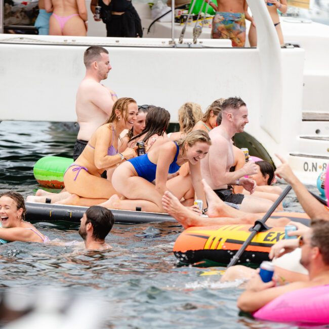A group of people are enjoying a sunny day on the water, gathered around a boat. Some are sitting on inflatables while others swim and chat. The scene is lively with colorful floats, including a pink flamingo. It’s an ideal setting akin to the vibrant atmosphere of Luxury Yacht Rentals Sydney or Boat Parties Sydney from The Yacht Social Club.