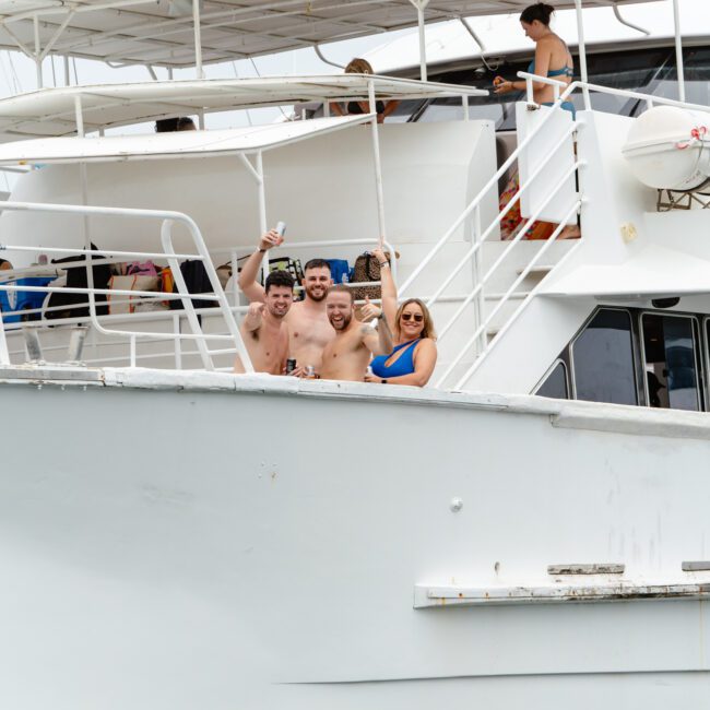 A group of people enjoying themselves on a white boat. Four are on the lower deck, three men and one woman, smiling and holding drinks. Above them, a woman stands on the upper deck. The water is calm, and the sky is overcast. Experience this with The Yacht Social Club Sydney Boat Hire.