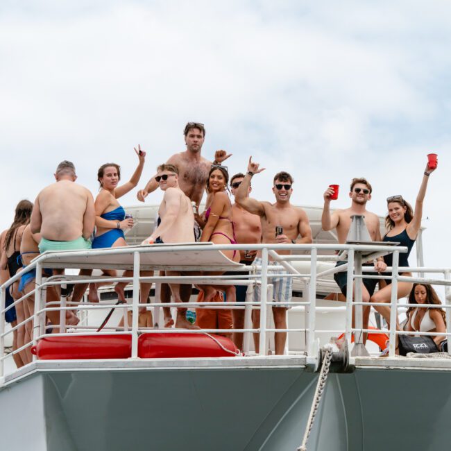 A group of people in swimwear are enjoying a party on The Yacht Social Club. They are standing on an upper deck, smiling, holding cups, and making celebratory gestures. The sky is cloudy, and the water is visible in the background.